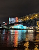 A brightly lit casino with neon signs and red lights is reflected in a river. A bridge stretches across the water, with an urban skyline visible in the background against an overcast night sky.