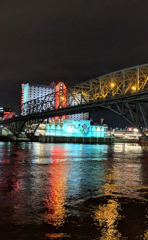 A brightly lit casino with neon signs and red lights is reflected in a river. A bridge stretches across the water, with an urban skyline visible in the background against an overcast night sky.