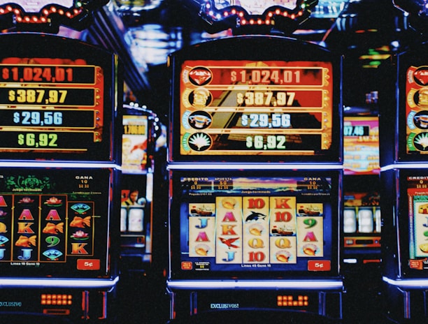 A row of colorful slot machines lit with bright neon lights, showcasing various graphical elements on the screens, indicating different games and jackpots. The atmosphere suggests a lively casino setting, bustling with excitement.