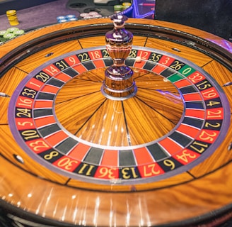 A roulette wheel with alternating red and black numbered pockets on a wooden platform. The wheel contains a small white ball resting in one of the pockets, indicating gameplay. Surrounding the wheel are stacks of colorful poker chips, suggesting a casino environment.
